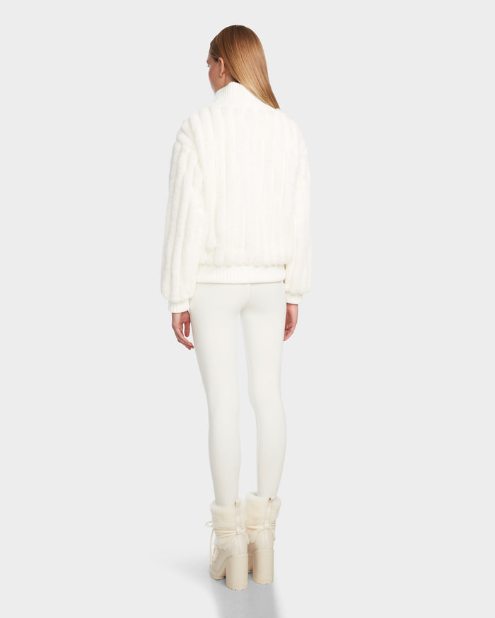 A person with long straight hair, facing away, wears the LAYLA - 8524610 Iceland women’s faux fur turtleneck jacket by Rudsak, paired with fitted white pants and white platform boots with fur details against a light background.
