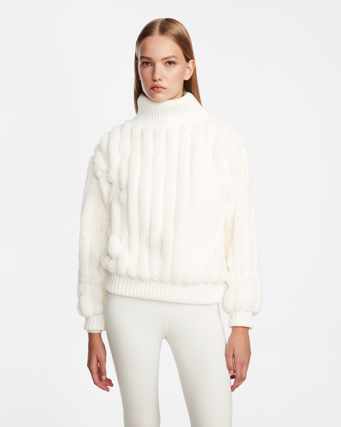A woman with straight, light brown hair wears the Rudsak LAYLA - 8524610 Iceland luxury white ribbed turtleneck sweater and matching pants, standing against a plain white background with a neutral expression and her arms by her sides.