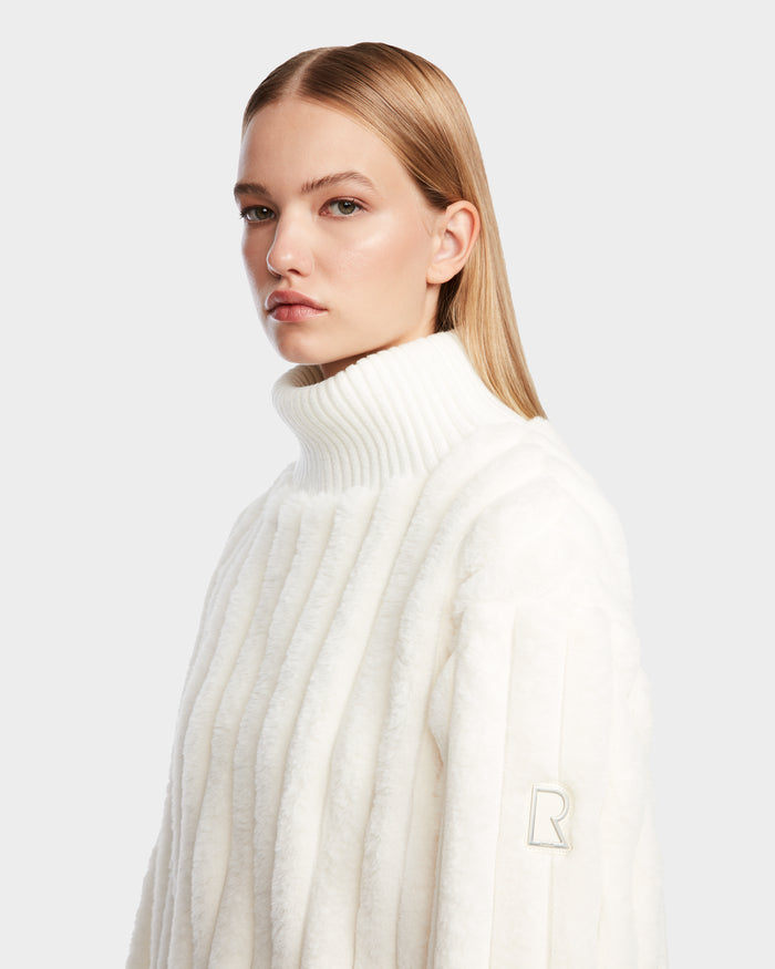 A woman with straight, blonde hair wears the LAYLA - 8524610 Iceland turtleneck sweater by Rudsak, featuring an R emblem on the sleeve, against a plain light background, gazing slightly to the side.