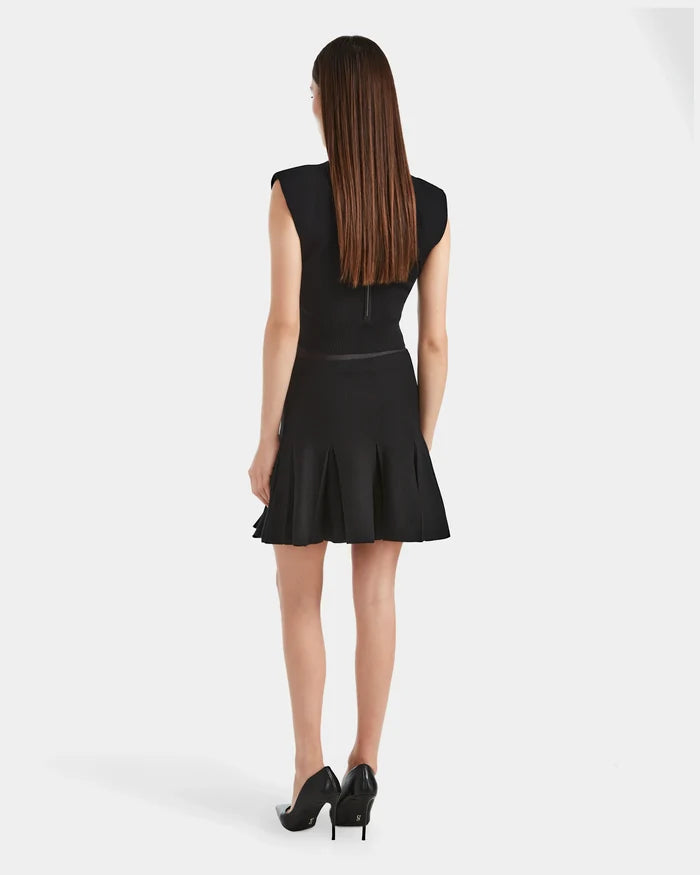A woman with straight brown hair wears the Rudsak LEA - 8524064 Black sleeveless dress with a high-waisted, flared pleated skirt and black heels, standing and facing away against a plain white background.