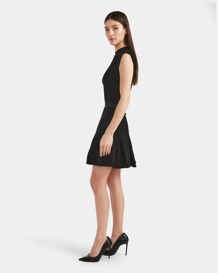 A woman with long brown hair in a sleeveless black dress and heels stands sideways against a white background, her look elevated by the Rudsak LEA - 8524064 Black high-waisted knit skirt.