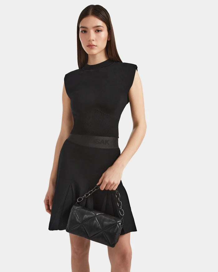 A woman with long brown hair wears the Rudsak LEA - 8524064 Black dress, featuring a contour soft knit top and pleated high-waisted skirt, holding a black quilted handbag with a chain strap against a white background.