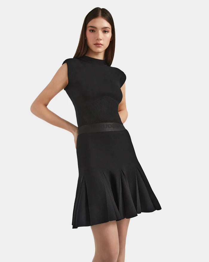A woman with long brown hair models the Rudsak LEA - 8524064 Black, an elegant sleeveless knit dress with a high-waisted pleated skirt, as she poses against a plain white background.