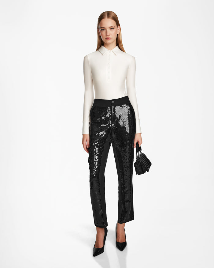 A woman poses against a white backdrop wearing black sequin trousers, a fitted white long-sleeve top, black pointed heels, and holding the Rudsak LEAR - 8524553 Black handbag.