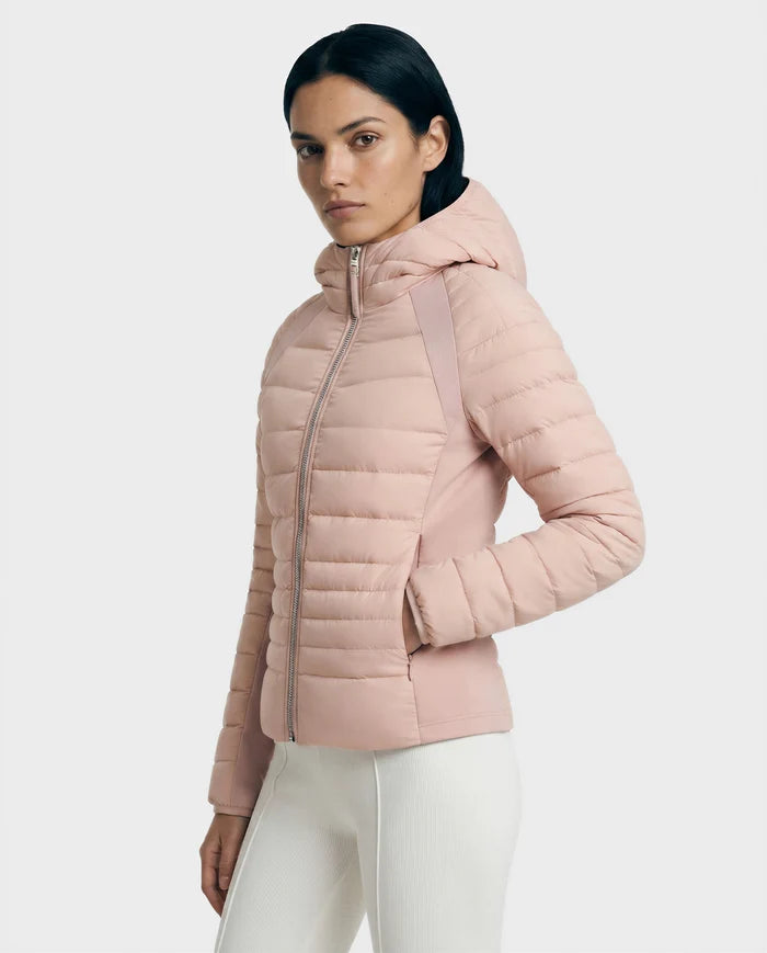 A woman with straight dark hair is wearing the Rudsak LENA - L124005 Soft Pink quilted jacket and white pants, standing against a plain light gray background and looking slightly to the side.