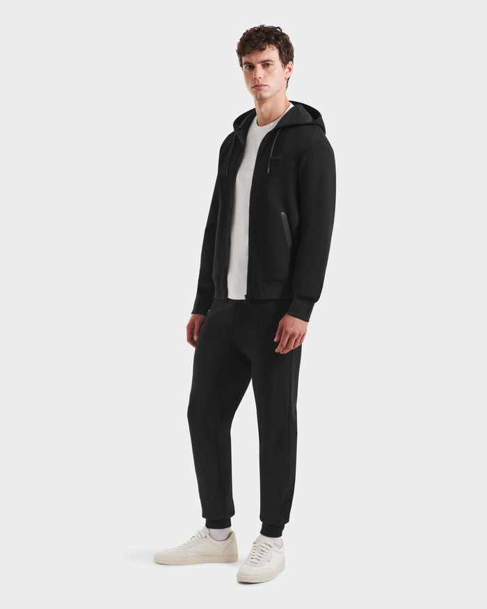 A man faces forward against a plain light background, wearing the Rudsak LEVI - 9525552 Black M unisex zip-up hoodie in premium cotton blend with vegan leather trims, paired with black joggers, a white tee, and white sneakers.