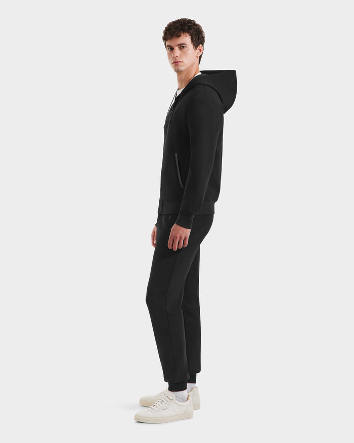 A man stands sideways wearing a Rudsak LEVI - 9525552 Black M hoodie, paired with matching black joggers and white sneakers against a plain light gray background.