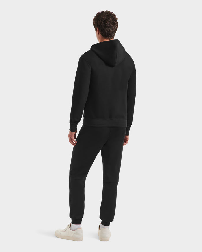 A person faces away from the camera, wearing the Rudsak LEVI - 9525552 Black M unisex zip-up hoodie with vegan leather trims and black jogger pants, paired with white sneakers, against a plain light background.