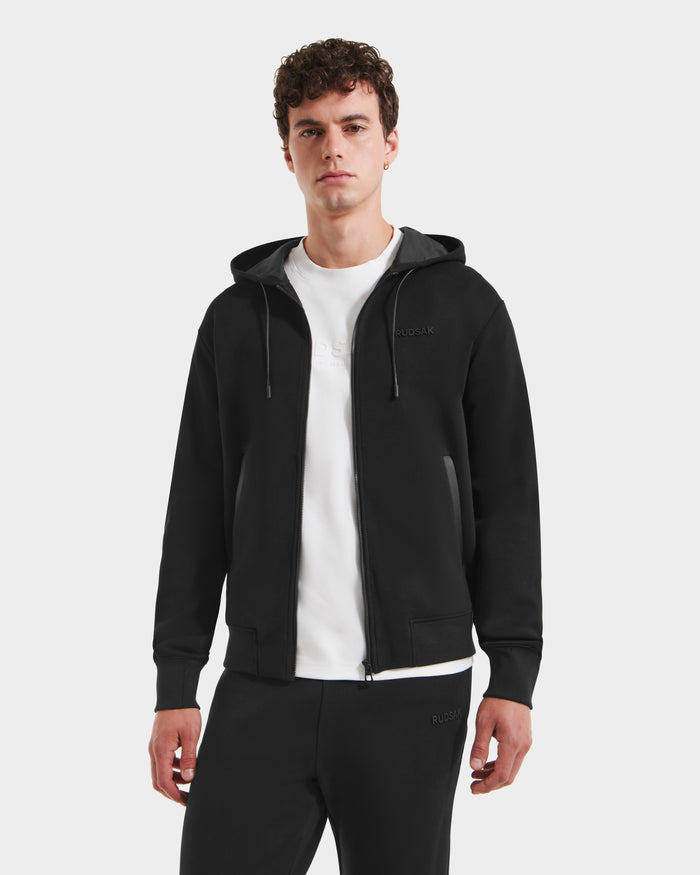 A person in the Rudsak LEVI - 9525552 Black M zip-up hoodie with vegan leather trims and matching black pants stands facing forward against a white background, showcasing elevated basics with effortless style.