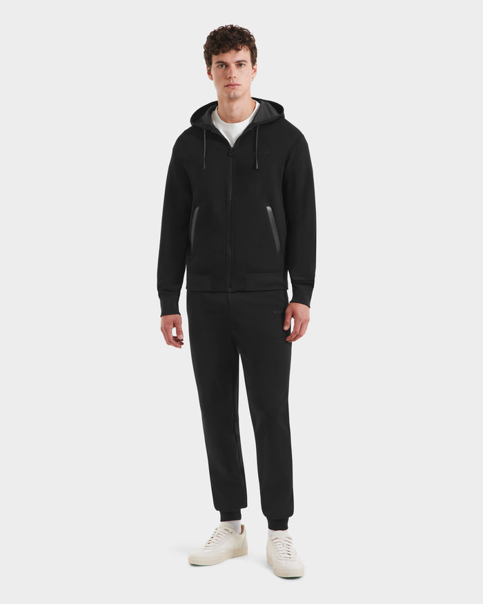 A man faces forward against a plain white background wearing the Rudsak LEVI - 9525552 Black M unisex zip-up hoodie with vegan leather trims, paired with matching black jogger pants, a white t-shirt, and white sneakers.