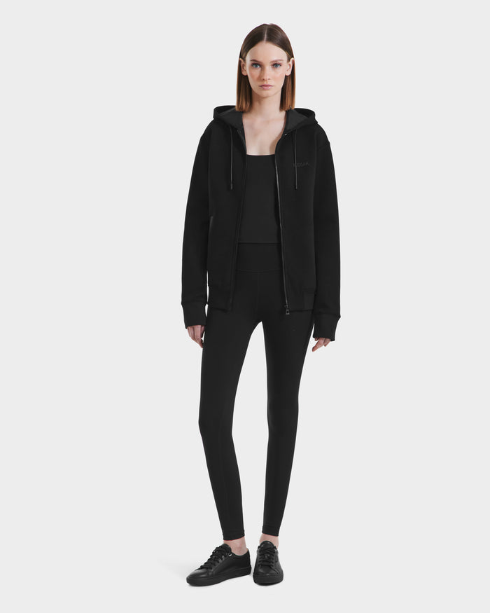 A woman faces forward against a white background, wearing the Rudsak LEVI - 9525552 Black W unisex zip-up hoodie with vegan leather trims, paired with a black tank top, leggings, and sneakers.