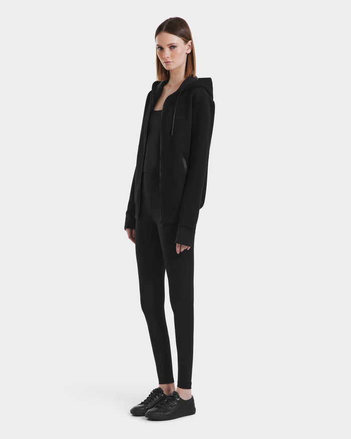 A woman with straight, shoulder-length hair and a neutral expression stands against a white background wearing the Rudsak LEVI - 9525552 Black W unisex zip-up hoodie with vegan leather trims, paired with a fitted top, leggings, and sneakers.