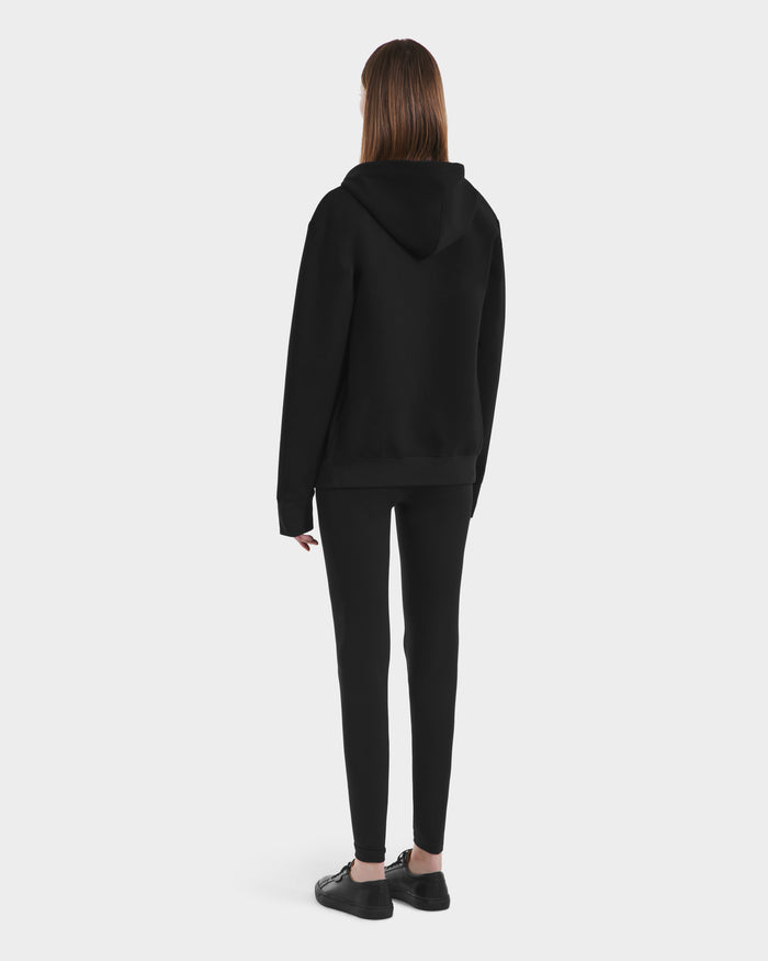 A person with straight, shoulder-length brown hair is shown from behind in a Rudsak LEVI - 9525552 Black W unisex zip-up hoodie with vegan leather trims, black leggings, and black shoes against a plain white background.