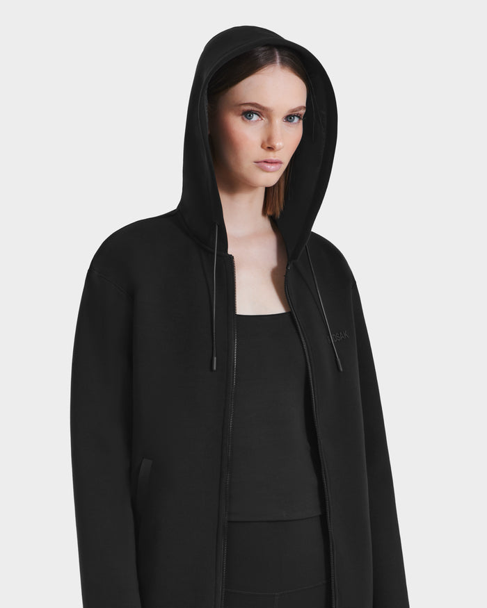 A woman with straight brown hair wears the Rudsak LEVI - 9525552 Black W unisex zip-up hoodie, featuring vegan leather trims, over a black top and looks slightly to the side against a plain white background.