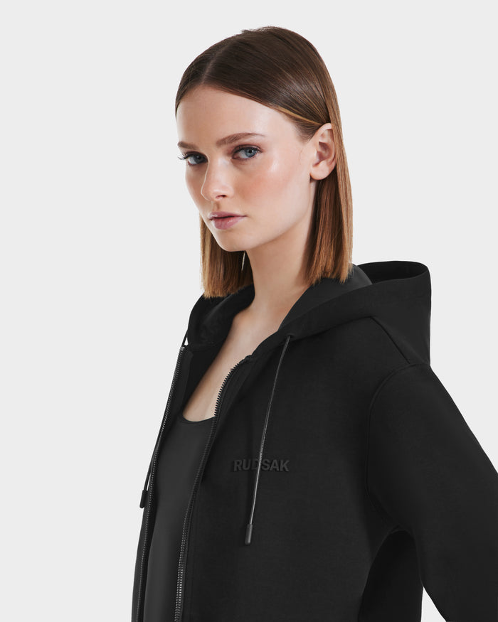 A woman with straight, shoulder-length brown hair wears the Rudsak LEVI - 9525552 Black W unisex zip-up hoodie with vegan leather trims and drawstrings over a black top, standing against a plain white background.