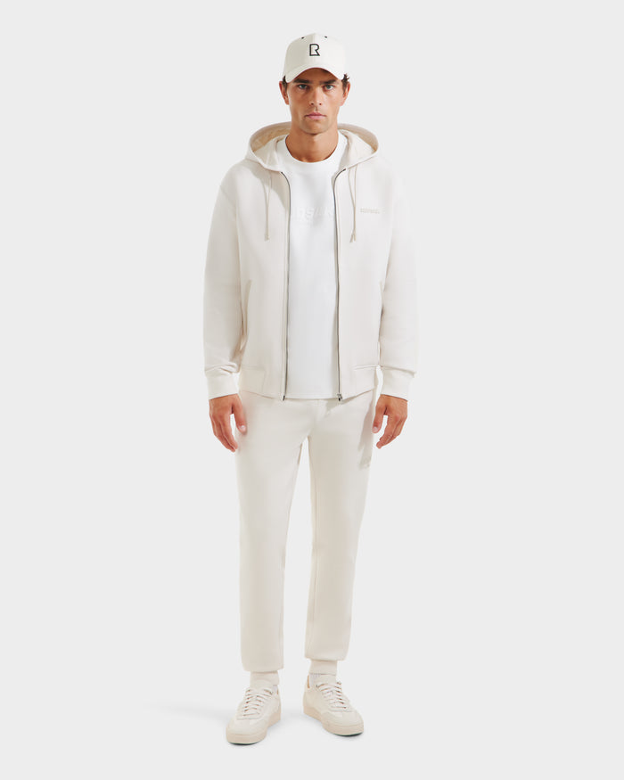 A man faces forward in a Rudsak LEVI - 9525552 Vanilla M unisex zip-up hoodie, t-shirt, jogger pants, sneakers, and a baseball cap—all white—set against a plain white background.