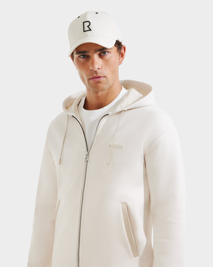 A man wears the Rudsak LEVI - 9525552 Vanilla M unisex zip-up hoodie, styled with a white T-shirt and a white cap featuring a black R logo, standing against a plain light background and looking at the camera.