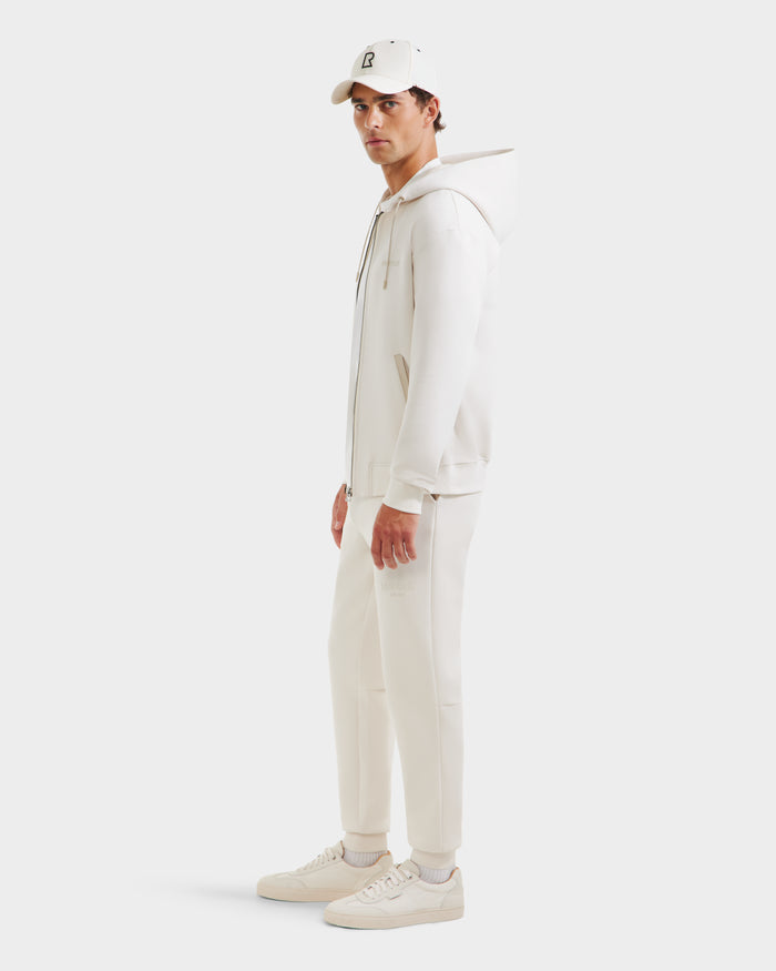 A man stands sideways in Rudsak’s LEVI - 9525552 Vanilla M: an off-white unisex zip-up hoodie with vegan leather trims, styled minimally with matching joggers, sneakers, and a cap against a plain light background.
