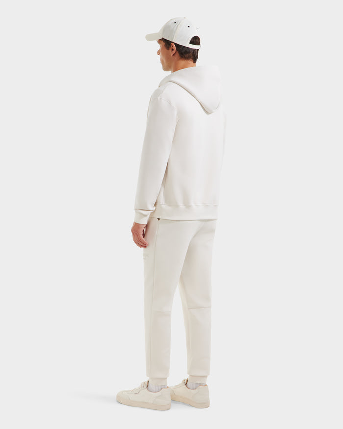 A man stands facing away, wearing the Rudsak LEVI - 9525552 Vanilla M unisex zip-up hoodie with vegan leather trims, paired with sweatpants, sneakers, and a baseball cap against a plain white background.