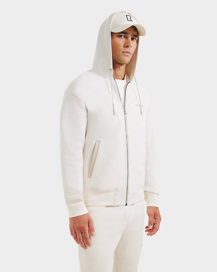 A man stands against a plain white background wearing the Rudsak LEVI - 9525552 Vanilla M unisex off-white zip-up hoodie with vegan leather trims, matching joggers, and a light-colored baseball cap.