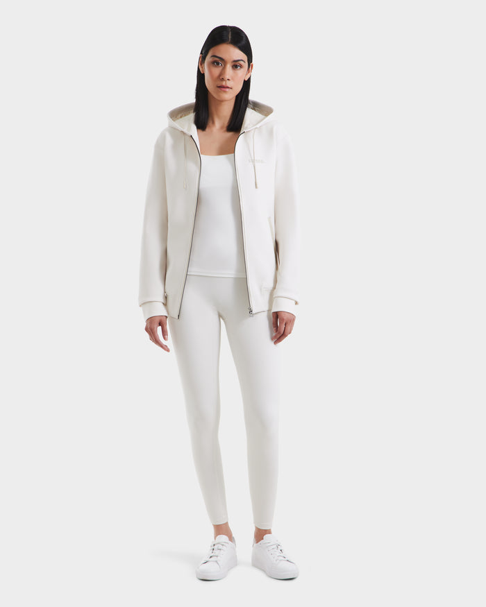 A woman with straight dark hair wears the Rudsak LEVI - 9525552 Vanilla W white zip-up hoodie with vegan leather trims, a white tank top, leggings, and sneakers, standing against a plain white background.