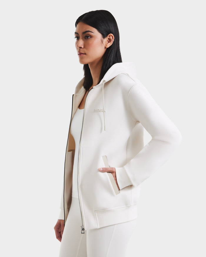 A woman with straight dark hair wears the Rudsak LEVI - 9525552 Vanilla W unisex zip-up hoodie and matching pants with vegan leather trims, standing with one hand in her pocket against a plain white background.