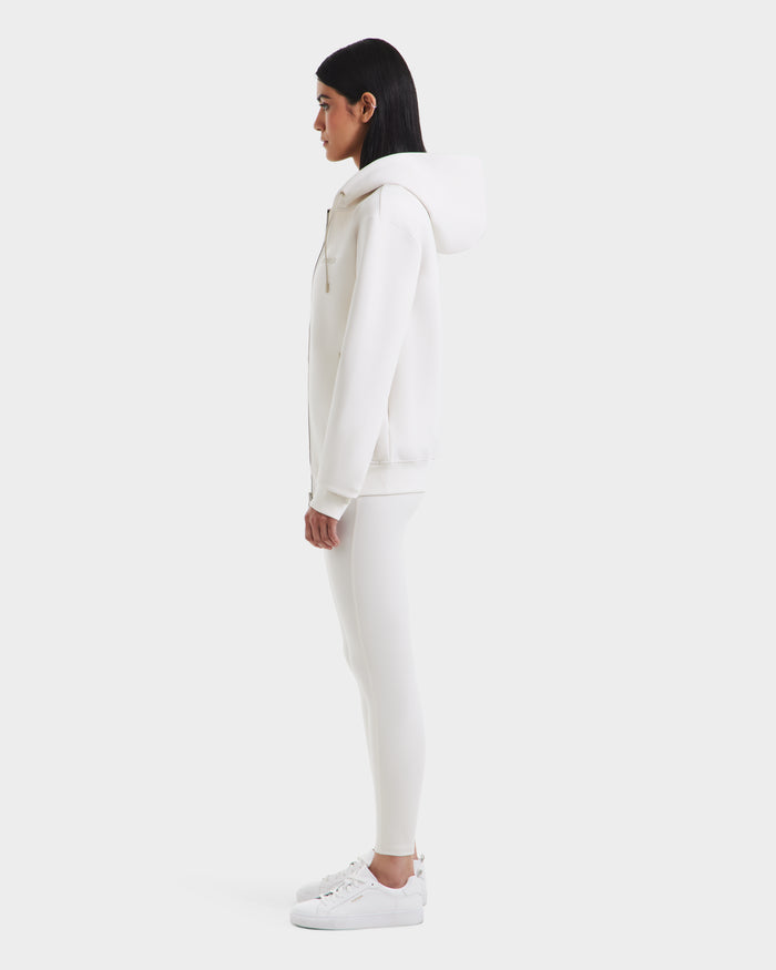 A woman stands in profile wearing the Rudsak LEVI - 9525552 Vanilla W ensemble: a hooded sweatshirt with vegan leather trims, matching leggings, and sneakers, all in white against a plain background.