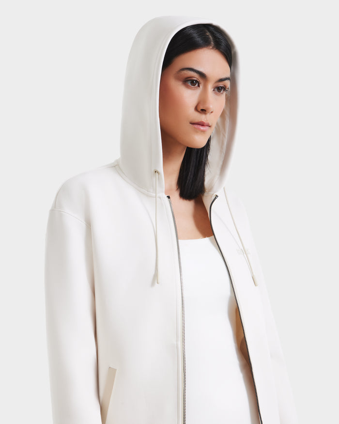 A woman with straight dark hair wears the Rudsak LEVI - 9525552 Vanilla W zip-up hoodie, featuring vegan leather trims, over a white top. The hood is up and the jacket partially zipped as she stands against a plain light background.