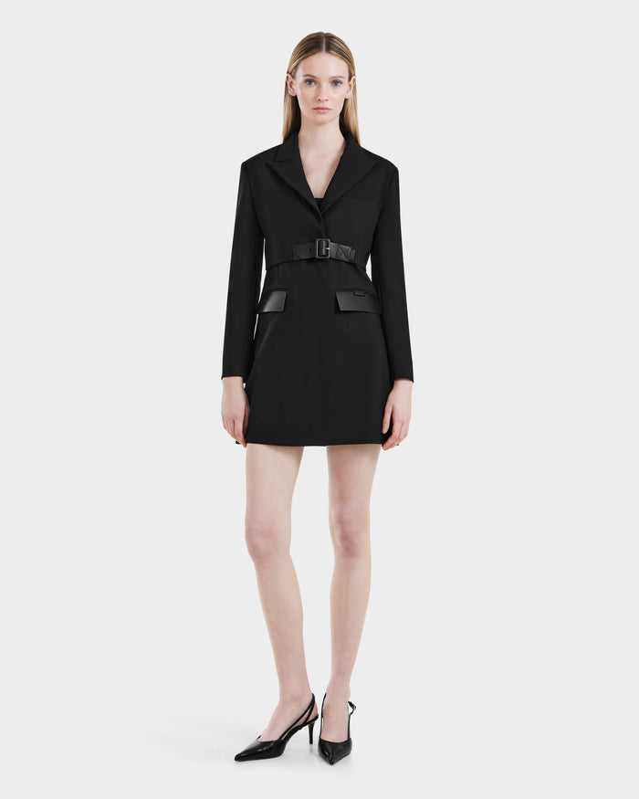 A woman faces forward in the Rudsak LEXI - 8525026 Black belted blazer dress with long sleeves and flap pockets, styled with a vegan leather belt and black pointed-toe heels, embodying modern power dressing against a white backdrop.
