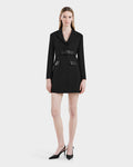 A woman faces forward in the Rudsak LEXI - 8525026 Black belted blazer dress with long sleeves and flap pockets, styled with a vegan leather belt and black pointed-toe heels, embodying modern power dressing against a white backdrop.
