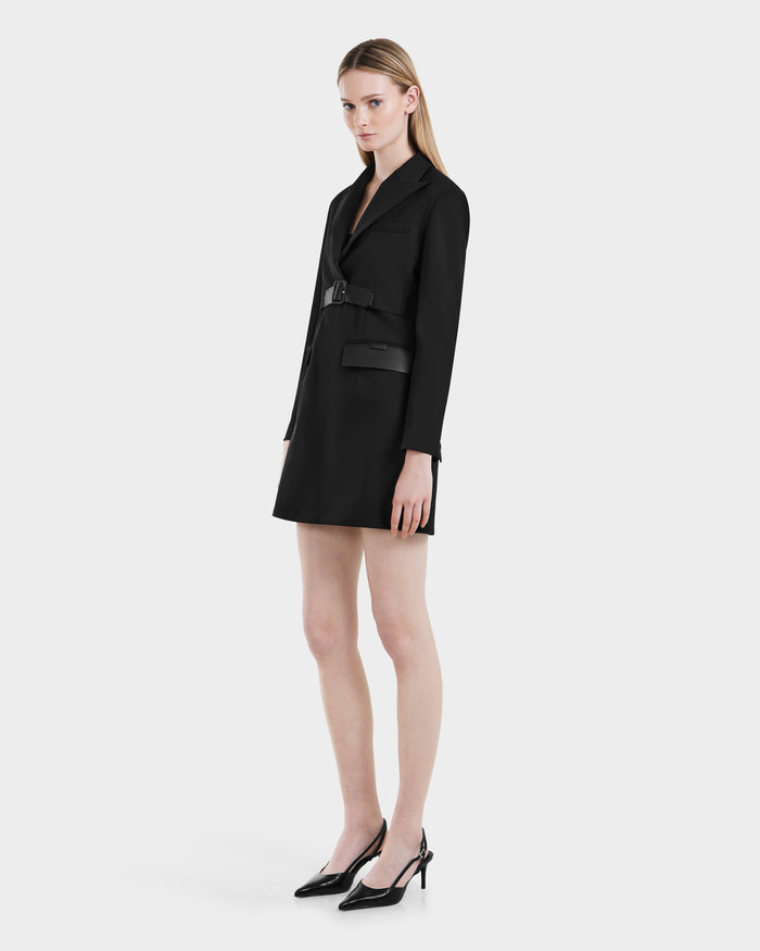 A woman with straight blonde hair and a neutral expression wears the Rudsak LEXI - 8525026 Black blazer dress, styled with a vegan leather belt and black pointed heels, standing against a plain light background.