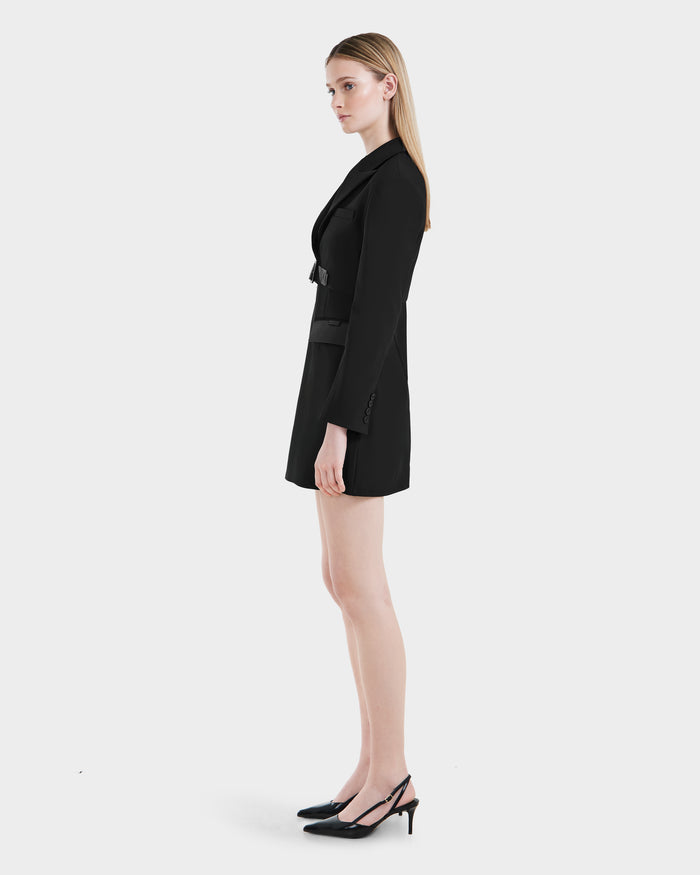 A woman with straight blonde hair stands in profile against a white background, wearing Rudsaks LEXI - 8525026 Black long-sleeve blazer dress with a vegan leather belt and black slingback heels for a power dressing look.