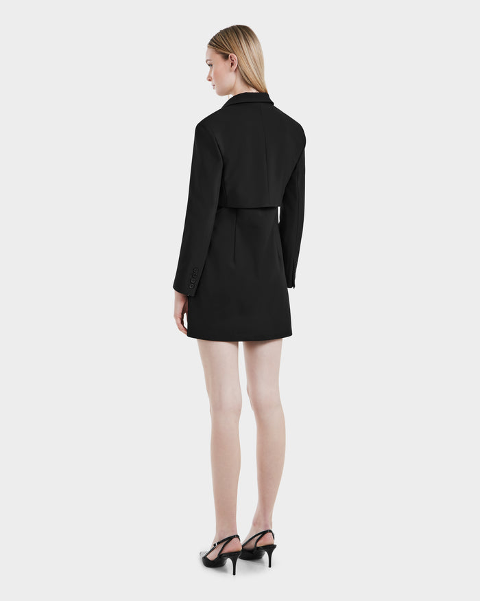 A woman exudes power dressing, standing with her back to the camera in the Rudsak LEXI - 8525026 Black blazer-style dress, cinched with a vegan leather belt and paired with black heels against a light gray background.
