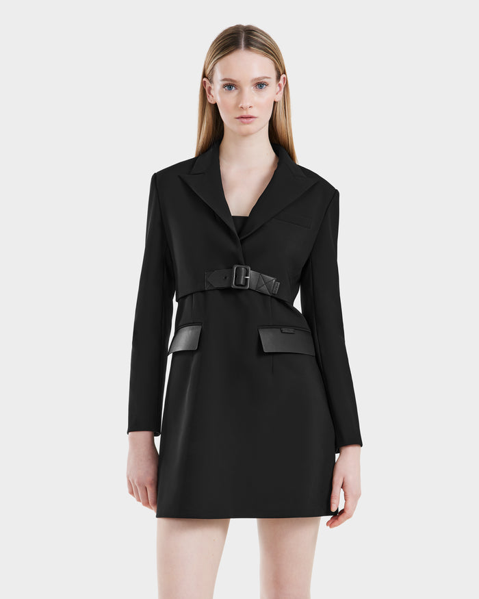 A woman with straight, long blonde hair wears the Rudsak LEXI - 8525026 Black tailored blazer dress, cinched with a vegan leather belt, standing against a plain light gray background for a chic power dressing look.
