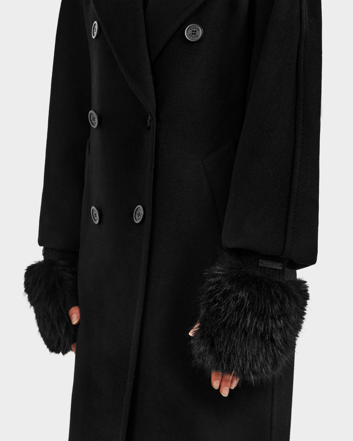 A person wears the Rudsak LIA - 8425007 Black long double-breasted coat with black faux fur cuffs and women’s fingerless mittens, their hands partly visible below the cuffs, standing against a plain white background.