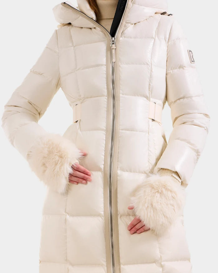 A person wears the Rudsak LIA - 8425007 Vanilla, a cream quilted puffer coat with hood, faux fur cuffs, front zipper, and high neck, standing with hands lightly touching at the waist.