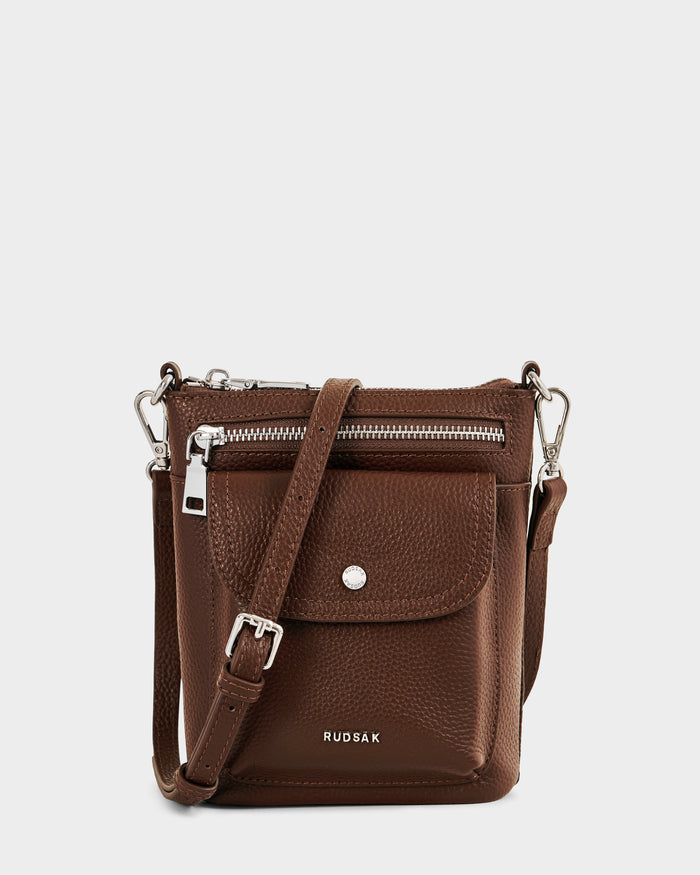 The Rudsak LILA - L325513 Mocha is a small brown women’s leather crossbody bag with a front pocket, silver zipper, button closure, adjustable strap, and RUDSAK logo at the bottom, shown on a plain white background.