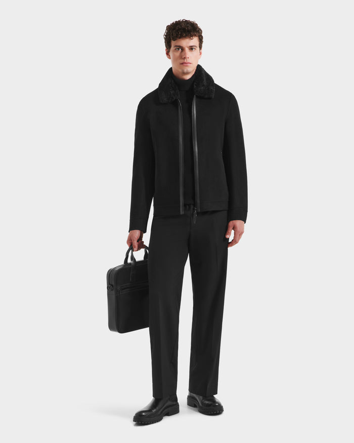 A man stands against a plain background wearing the Rudsak LINCOLN - 6125038 Black mens wool coat with a removable faux fur collar, paired with black clothes and shoes. He holds a black briefcase in his left hand, looking straight ahead.