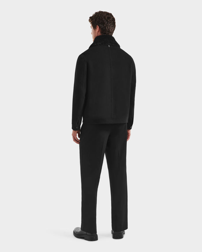 A man stands facing away, wearing the Rudsak LINCOLN - 6125038 Black mens wool coat with removable faux fur collar, paired with black trousers and shoes against a plain light background.