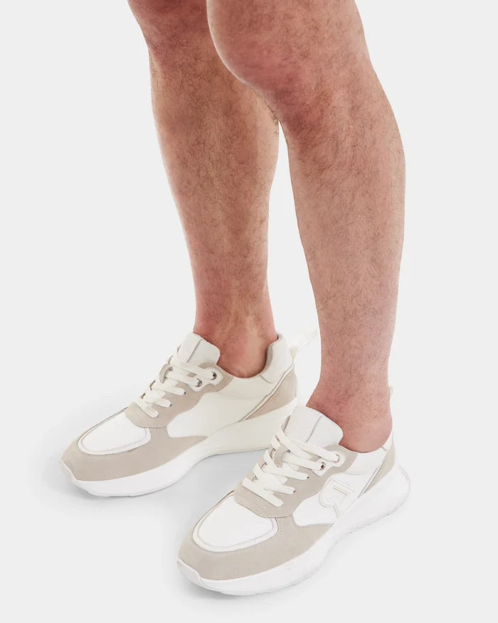 A person wearing Rudsak LIONEL - 6225001 Iceland men’s white and beige lace-up sneakers with a modern design, standing on a white background; only their lower legs and feet are visible.