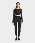 A woman stands facing forward, wearing the Rudsak LORA - 8524026 black activewear top with high-waisted black leggings and black and white sneakers, set against a plain light background.
