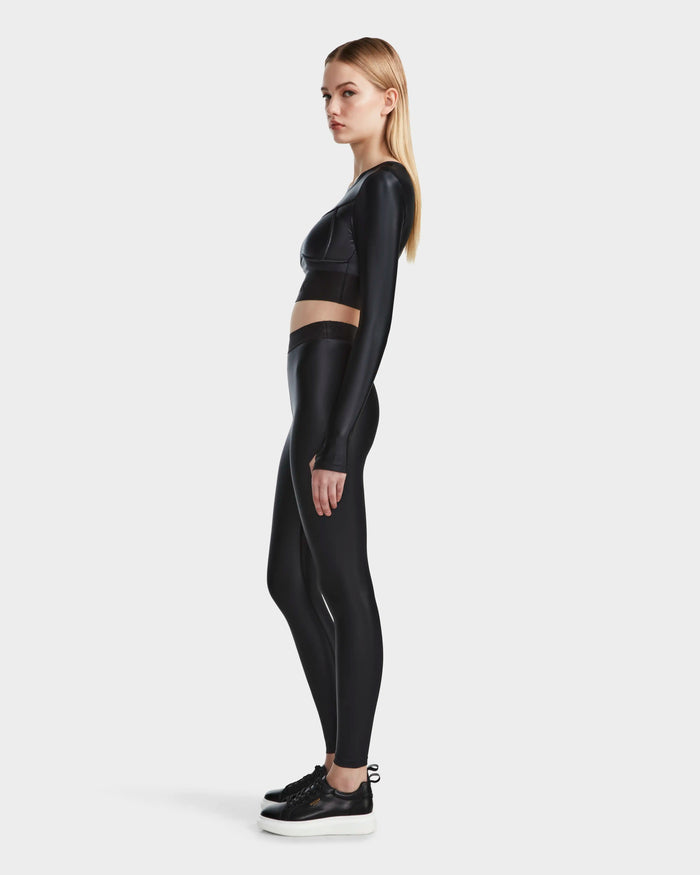 A woman stands in profile against a plain white background wearing Rudsak’s LORA - 8524026 Black long-sleeve activewear top, high-waisted black leggings, and black sneakers.