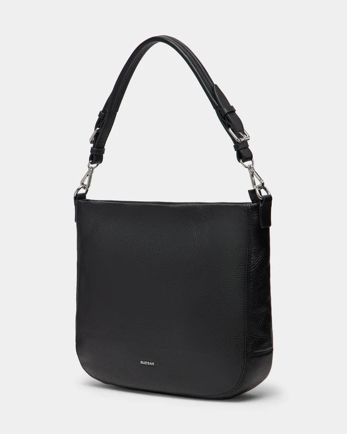 The Rudsak LUCYLE - 8325012 Black is a medium-sized, curved rectangular leather hobo bag with silver hardware, a wide adjustable strap, and subtle embossed branding near the bottom center, shown on a plain white background.