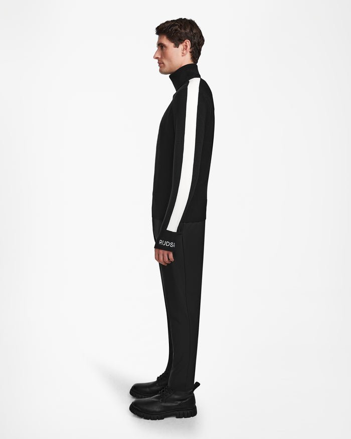 A man in profile against a white background wears the Rudsak LUKAS - 6524543 Black mens knit turtleneck sweater with a bold white sleeve stripe—ideal for après-ski or winter sports-inspired looks.