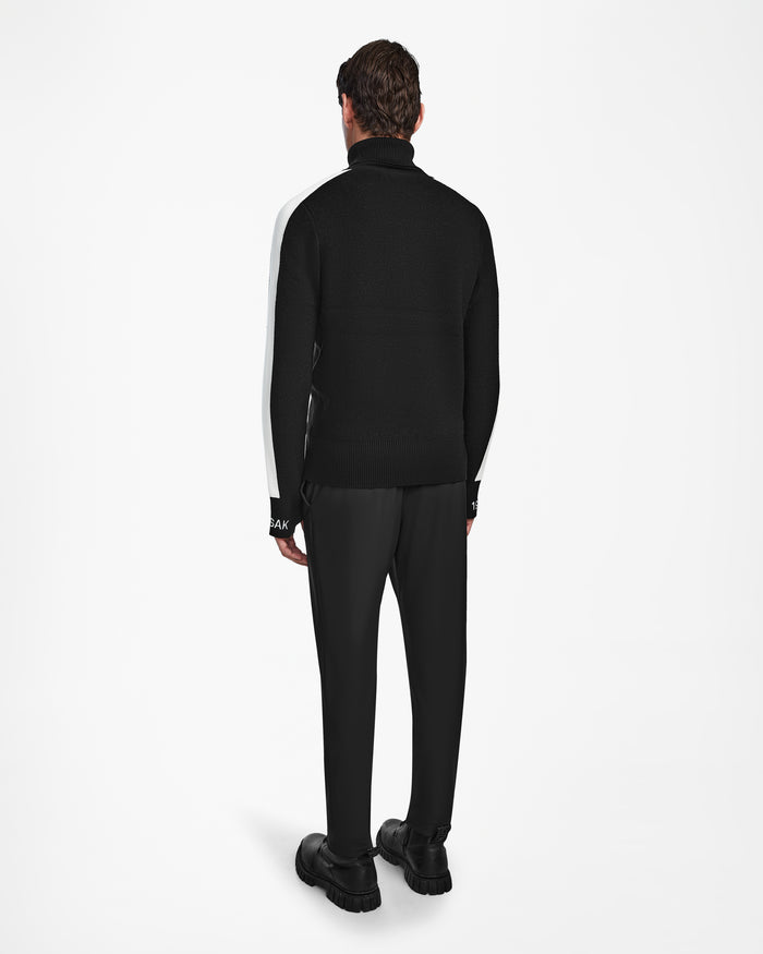A man faces away from the camera, styled in Rudsaks LUKAS - 6524543 Black jacket over a men’s knit turtleneck, with black pants and chunky shoes—an effortlessly cool look against a plain white background.