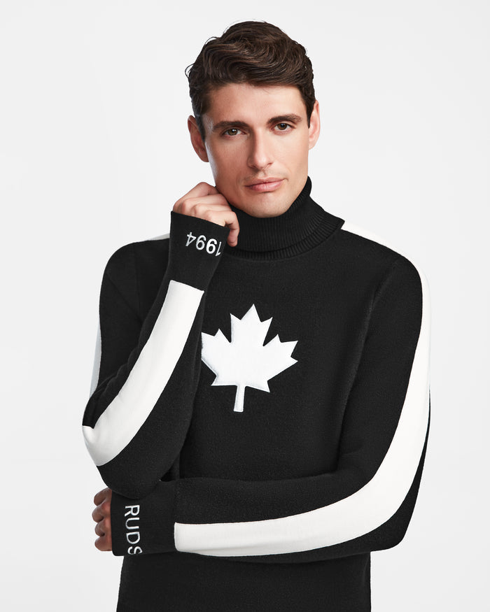 A man with short brown hair models the Rudsak LUKAS - 6524543 Black men’s knit turtleneck après-ski sweater featuring a white maple leaf on the chest and white sleeve stripes, posing against a plain white background.