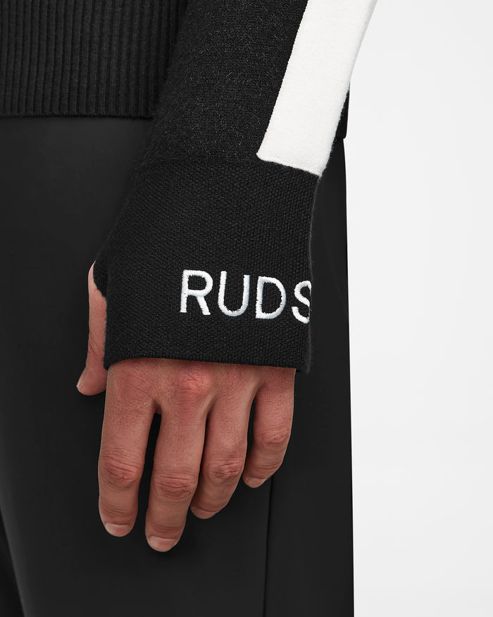 A close-up of a person’s left hand in Rudsak’s LUKAS - 6524543 Black men’s knit turtleneck sweater, featuring “RUDSAK” embroidered in white on the sleeve and paired with black pants—ideal for après-ski style.