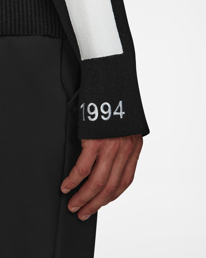 A close-up of a person’s arm wearing the Rudsak LUKAS - 6524543 Black men’s knit turtleneck sweater, featuring a white stripe and “1994” embroidery near the cuff, evokes classic après-ski style against a plain white background.