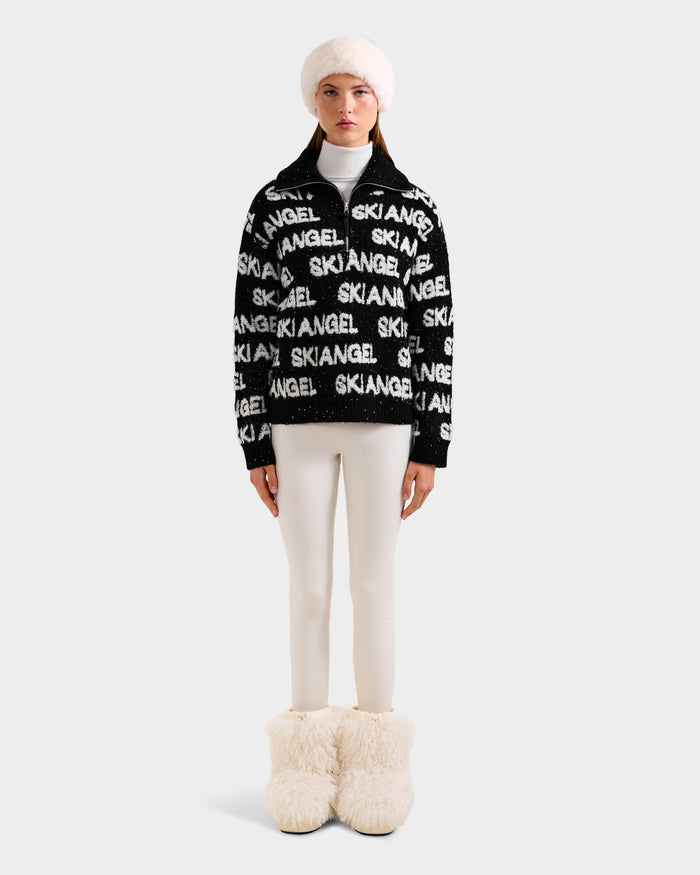 A person stands facing forward in the Rudsak LYDIA - 8525687 Black women’s ski sweater with white SKI ANGEL text, paired with white fitted pants, fluffy white boots, and a white fur hat against a plain light background.