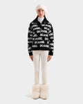 A person stands facing forward in the Rudsak LYDIA - 8525687 Black women’s ski sweater with white SKI ANGEL text, paired with white fitted pants, fluffy white boots, and a white fur hat against a plain light background.